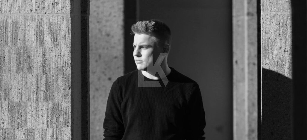 A young man wearing a dark shirt stands between concrete pillars, looking to the left. Sunlight casts strong shadows, creating a dramatic black-and-white effect.