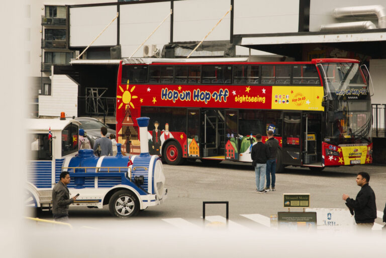 A colorful Hop-On Hop-Off Ålesund sightseeing bus is parked beside a small blue and white tourist train. Several people stand nearby in an urban setting with buildings in the background in Ålesund, Norway.