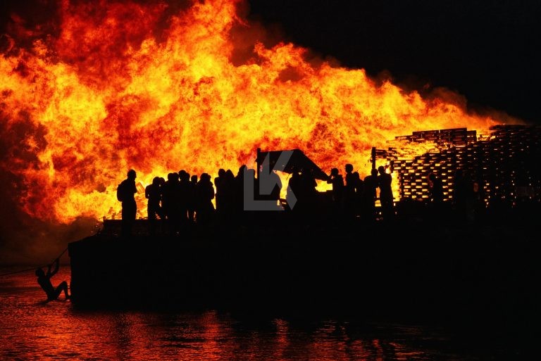 Silhouetted figures stand before the massive, blazing Slinningsbålet 2026 bonfire at night, with one person pulling a rope in the water in the foreground.