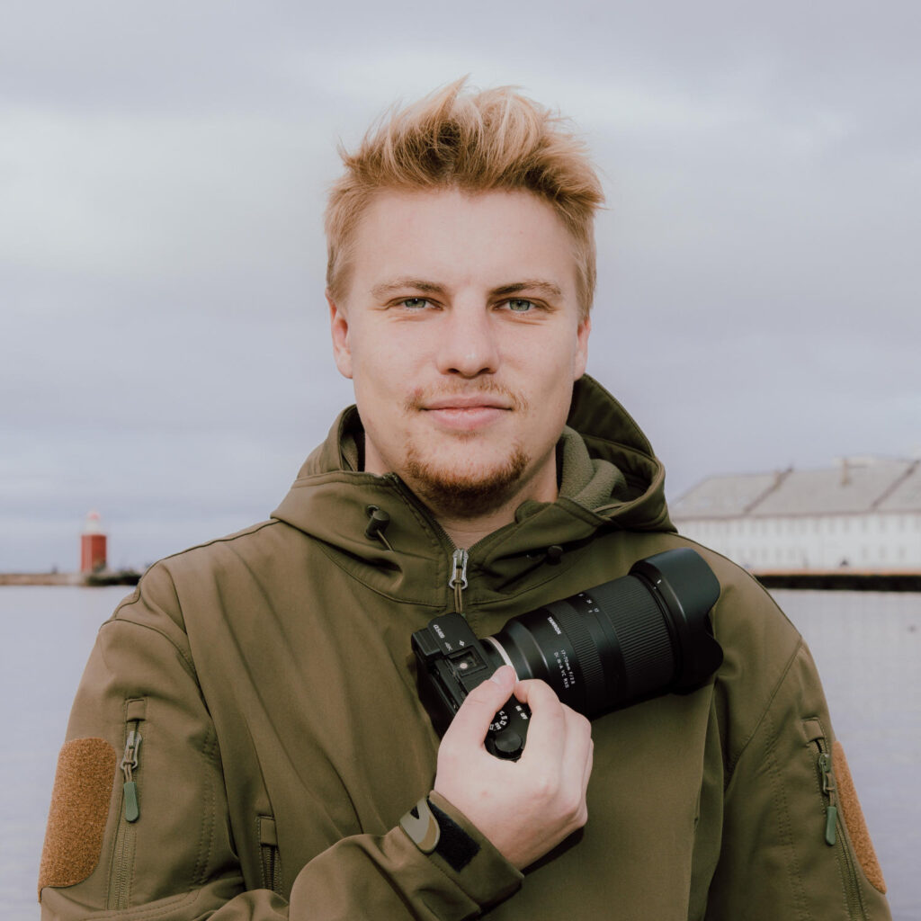 A young man with blond hair, wearing an olive green jacket, stands by the water holding a camera. A lighthouse and a white building are visible in the background under a cloudy sky in Norway, capturing Lukas Klein’s adventurous spirit.